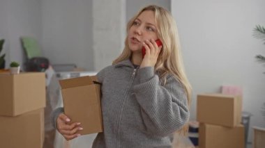 Oturma odasında elinde kutuyla telefonla konuşan bir kadın, yeni evdeki kutuları taşıyor, sarı saçlı, rahat, rahat, kapalı bir ortamda..