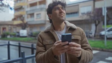 İspanyol bir adam şehir sokağında akıllı telefon kullanıyor. Üzerinde kahverengi bir ceket, arka planda park edilmiş arabalar ve binalar var..