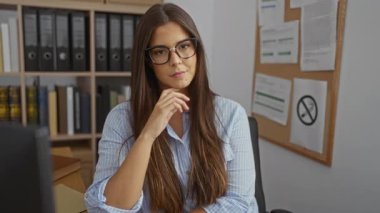 Ofisteki genç bir kadın, düşünceli bir yüz ifadesi ve uzun esmer saçı var. Arka planda dosyaları ve ilan panosuyla bir masada oturuyor.