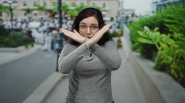 Yoğun bir caddede çapraz ellerle el kol hareketi yapan bir kadın, canlı bir şehir çevresiyle çevrili, ciddi bir ifadeyle reddedilmeyi reddediyor..