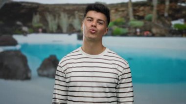 Çizgili gömlekli genç bir adam tatil köyü havuzunun yanında neşeli bir poz veriyor, otel ortamının boş havasını büyüleyici bir ifadeyle sergiliyor..