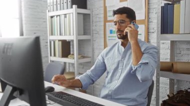 Modern iş yerindeki ofis masasında sakallı genç adam konuşuyor rafları ve bilgisayarı var..