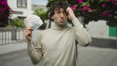 Şehir caddesinde paralar tutan adam şaşırmış bir ifadeyle, canlı şehir manzarası ve renkli çiçeklerle çevrili, açık havada finansal başarıyı gösteren.