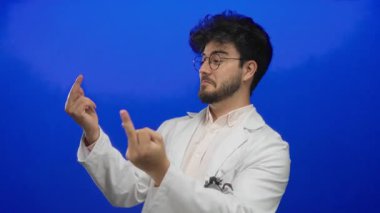 Sakallı, beyaz önlüklü genç adam orta parmaklarıyla mavi bir arka plana doğru esprili hareketler yapıyor. Gülümsüyor ve neşeli ama isyankar bir tavır sergiliyor..