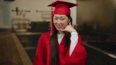 Kırmızı mezuniyet kıyafeti giymiş bir kadın mutfakta gülümsüyor, bir telefon jesti yapıyor, ev ortamındaki başarıyı somutlaştırıyor..