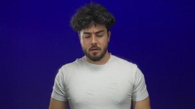 Sakallı genç adam mavi arka planda hapşırıyor, gözlerini kapatıyor ve beyaz gömlek giyerken burnunu tutuyor, rahatsızlığı ve soğuk algınlığı belirtilerini gösteriyor, İspanyol erkek sağlığı.