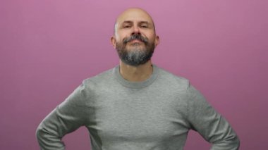 Kel kafalı sakallı adam güvenli bir şekilde pembe arka plana karşı duruyor. Pozitif ve ulaşılabilir bir hava yayıyor.