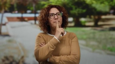 Gözlüklü İspanyol kadının güneşli bir parkta yaratıcı bir jesti var. Açık havada canlı bir yeşilliğin ortasında iç gözlem ve düşüncelilik sergiliyor..