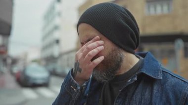 Şehir caddesinde kel sakallı bir adam. Yüzünde elleri, kot ceketi ve beresi var..