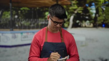 Gözlüklü genç İspanyol bir adam park ortamında notlar alıyor, kırmızı bir gömlek ve önlük giyiyor, açık havada düşünceli bir ifadeyle yakalanmış, çekici özelliklerini geliştiriyor..