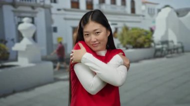 Kırmızı giyinmiş, açık havada gülümseyen, kendini sıcak tutan, güneşli bir sokakta mutluluk ve memnuniyet gösteren bir kadın..