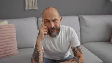 Oturma odasındaki koltukta oturan sakallı, beyaz tişörtlü, düşünceli, sakallı adam.