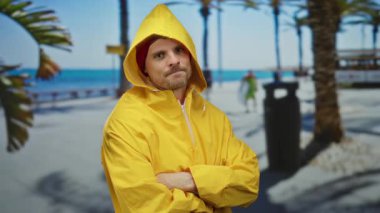 Sarı yağmurluklu genç adam kollarını kavuşturmuş güneşli sahil geçidinde palmiye ağaçları ve arka planda okyanusla geziyor..