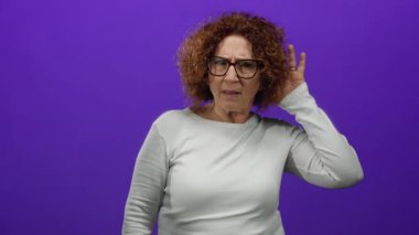 Gözlük ve beyaz gömlek giyen, göz kamaştırıcı mor bir arkaplan önünde el pençe divan duran kadın şaşkınlık ve şaşkınlık içinde..