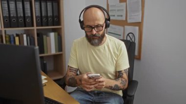 Ofiste kulaklık takan sakallı ve dövmeli İspanyol bir adam arka planda klasörleri olan bir masada otururken akıllı telefonuna odaklanmış..