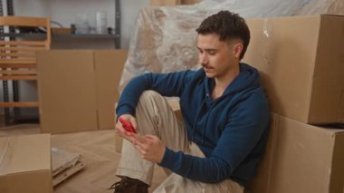 Yerde oturan genç adam yeni apartman dairesinde kutularla çevrili bir şekilde telefonla erkek gevşeme teknolojisini gösteriyor.