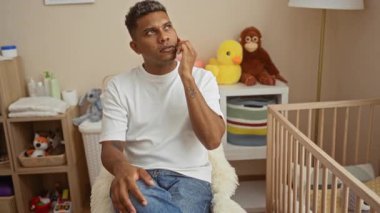 Rahat bir yatak odasında telefonla konuşan genç bir adam yumuşak oyuncaklarla çevrili bir bebek beşiğinin yanında oturmuş, rahat ve aile odaklı bir ev ortamı yaratıyor..