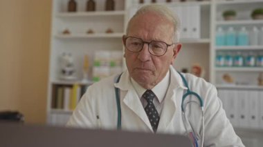 Üniformalı kıdemli erkek doktor, tıbbi ekipman ve malzeme raflarıyla çevrili bir hastane kliniğinde dizüstü bilgisayarla çalışırken dikkatle odaklanıyor..