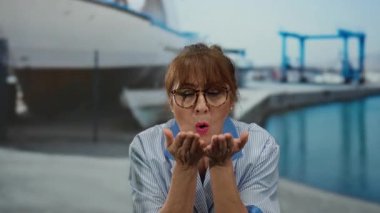 Üniformalı İspanyol kıdemli bir kadın deniz kıyısında teknelerle kameraya bir öpücük yolluyor. Açık havada çekilmiş sıcak ve davetkar bir sahne sergiliyor..