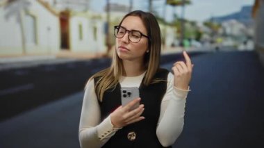 Güneşli bir sokakta elinde akıllı bir telefonla, gözlük takan ve günlük kıyafetler giyen bir kadın.