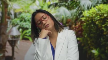 Beyaz ceketli ve kırmızı gözlüklü Çinli bir kadın canlı yeşil bir parkta dinleniyor. Genç zarafetini sergiliyor ve doğal güzelliğini sergiliyor..