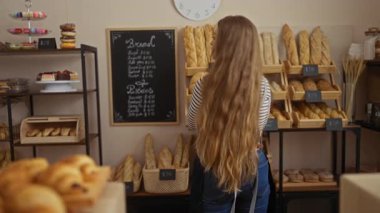 Fırında gülümseyen bir kadın, arka planda ekmek rafları, pastalar ve karatahta menüsüyle, çizgili gömlek ve önlük giyiyor.