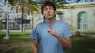 Parmaklı dudaklı bir adam güneşli yeşil bir parkta sessizlik işareti yapıyor..