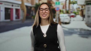 Kadın, enerjik bir şekilde, açık havada, gözlük ve siyah yelek takan, şehir sahnesine güven ve pozitiflik saçan, enerjik bir şekilde parmağıyla işaret ediyor..