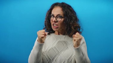 Kıvırcık saçlı ve gözlüklü bir kadın güçlü duygular gösteren bir süveterle canlı mavi bir arka plana karşı öfkesini ifade ediyor..