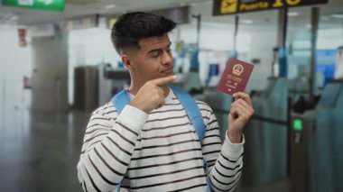 Çin pasaportu taşıyan bir adam havaalanı terminalinde parmaklarıyla kafaya dokunur. Uçuş işaretleri ve kapalı alanlarda seyahat için düşünceli bir hazırlık yakalar..