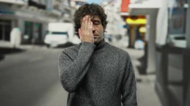 Bir şehir caddesinde İspanyol bir adam bir gözünü kapatıyor gri bir kazak giyerken stresli görünüyor, şehir stresi ve duygularını yakalıyor..