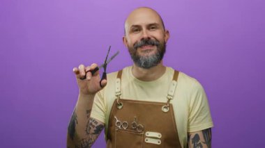 Önlük takan ve elinde makas tutan kel sakallı bir adam canlı mor bir arka plana, dövmeli kollara ve kuaför aletlerine güvenerek gülümsüyor..