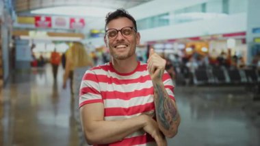 Yoğun bir havaalanı terminalinde sevinçten sırıtan çizgili tişörtlü genç adam, yolcularla çevrili seyahat planları ve canlı iç mekanlar hakkındaki heyecanını sergiliyor..