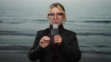 Plaj kıyısında mikrofonlu genç bir kadın muhabir açık gökyüzü altında deniz altlarında gözlük takıyor ve açık hava ortamında siyah ceket giyiyor..
