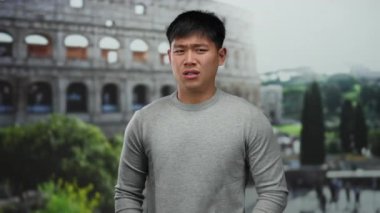 Gri kazaklı genç Asyalı adam yeşillik ve şehir hayatı ile çevrili ikonik Roma kolezyumunun önünde yüzünü buruşturarak samimi bir anı yakalıyor..