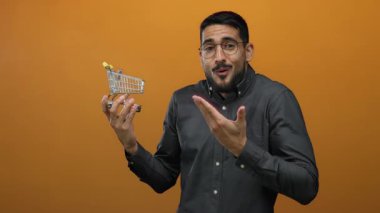 Canlı turuncu arka planda minyatür alışveriş arabası tutan genç adam, karışık duygular ifade ediyor, izole ve gündelik kıyafetlerle moda, online alışveriş temasını vurguluyor.