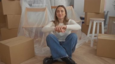 Yeni evinde gülümseyen bir kadın. Etrafı kutularla çevrili bir şekilde yerde oturuyor. Kapalı bir ortamda yeni bir başlangıç yapıyor..