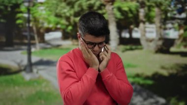 Kırmızı kazaklı genç adam yeşil park manzarasıyla çevrili bir akıllı telefondan konuşuyor..