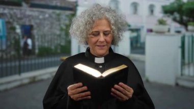Ruhban sınıfında gri saçlı yaşlı bir kadın sokak ortamında kitap okuyor..
