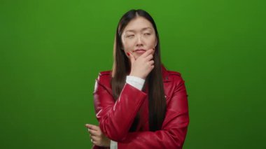 Kırmızı ceketli genç bir kadın yeşil arka plana kafa yoruyor, zarif ve kendine güvenen, elleri çenesinde düşünceli bir ifade takınıyor..