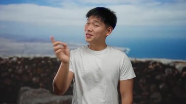 Genç Çinli bir adam arka planda okyanus manzaralı deniz kenarında neşeli bir jest yapıyor, beyaz bir gömlek giyiyor ve açık yürekli bir tavır sergiliyor.