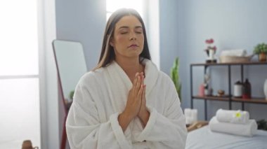 Kaplıca odasında meditasyon yapan, beyaz bir bornoz giyen, elleri dua ederken aynanın önünde duran ve sağlıkla ilgili şeylerle dolu rafları olan bir kadın.