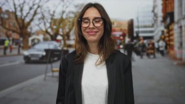 Londra 'da bir şehir caddesinde gülümseyen İspanyol kadın, gözlüklü ve beyaz gömlekli, şehir trafiği ve insanlarla çevrili..