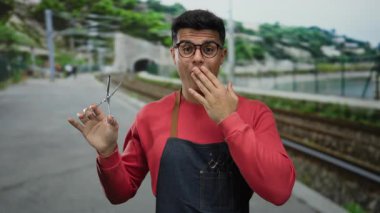 Genç Latin asıllı erkek kuaför deniz kenarında geziniyor, açık havada gözlük ve önlük takıyor, mesleğini canlı bir şekilde gösteriyor..