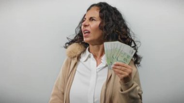 Orta yaşlı bir kadın beyaz arka planda dolarları tutuyor, gürültü rahatsızlığı nedeniyle kulakları örtüyor, elinde izole banknotlarla rahatsız edici şeyler söylüyor..