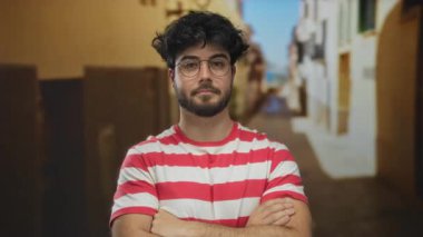 Sakallı ve gözlüklü genç adam kollarını açık bir sokakta çapraz yapıp kırmızı ve beyaz çizgili bir gömlek giyerek rahat ve rahat bir hava veriyor..