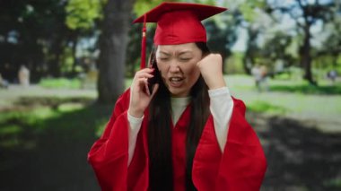 Mezuniyet elbisesi ve kepi giymiş bir kadın park ortamında telefonda hayal kırıklığını ifade ediyor, yemyeşil bir arka plana karşı öfke ve stresi somutlaştırıyor, yetişkin sorunlarını sembolize ediyor.