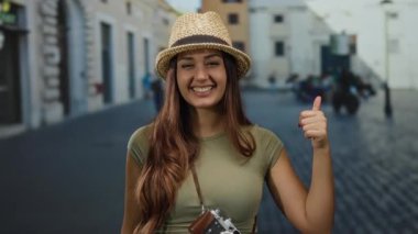 Güneşli caddede elinde kamerayla gülümseyen bir kadın başparmak kaldırıyor, neşeli keşifler yapıyor ve dışarıda kendine güveni olan olumlu seyahat titreşimleri yayıyor..