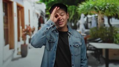 Genç adam güzel bir jest yapıyor, canlı şehir caddesinde, kot giyiyor, şehir yeşilliğinin ortasında neşeli bir ifade ve günlük kıyafetle mutluluk ve güven ifade ediyor..