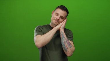Kolu dövmeli genç adam canlı yeşil arka planda poz veriyor, elleri yanağın altına sıkışmış uyku ya da rahatlama hissi uyandırıyor, günlük gömlek giyiyor, yakışıklı ve kendinden emin görünüyor..